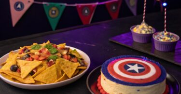 Un plateau de nachos avec des sauces colorées représentant les pierres de l'Infini, éclairées par une lumière violette. A coté Un cheesecake décoré en forme du bouclier de Captain America, entouré de petits drapeaux Avengers.