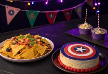 Un plateau de nachos avec des sauces colorées représentant les pierres de l'Infini, éclairées par une lumière violette. A coté Un cheesecake décoré en forme du bouclier de Captain America, entouré de petits drapeaux Avengers.